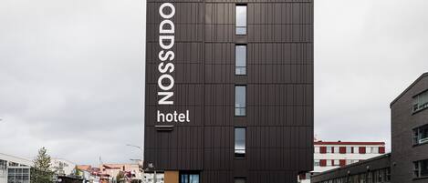 A modern hotel building with a vertical sign reading "NOSSADO hotel" and a visible entrance with steps leading up to it.