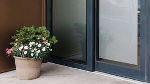 Entrada de un edificio con un gran escaparate de vidrio, una planta en maceta con flores y un logotipo visible en la puerta.