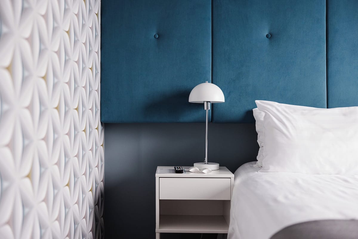 Una cama bien hecha con sábanas blancas, una mesita de noche blanca con una lámpara, y una pared con un diseño geométrico.