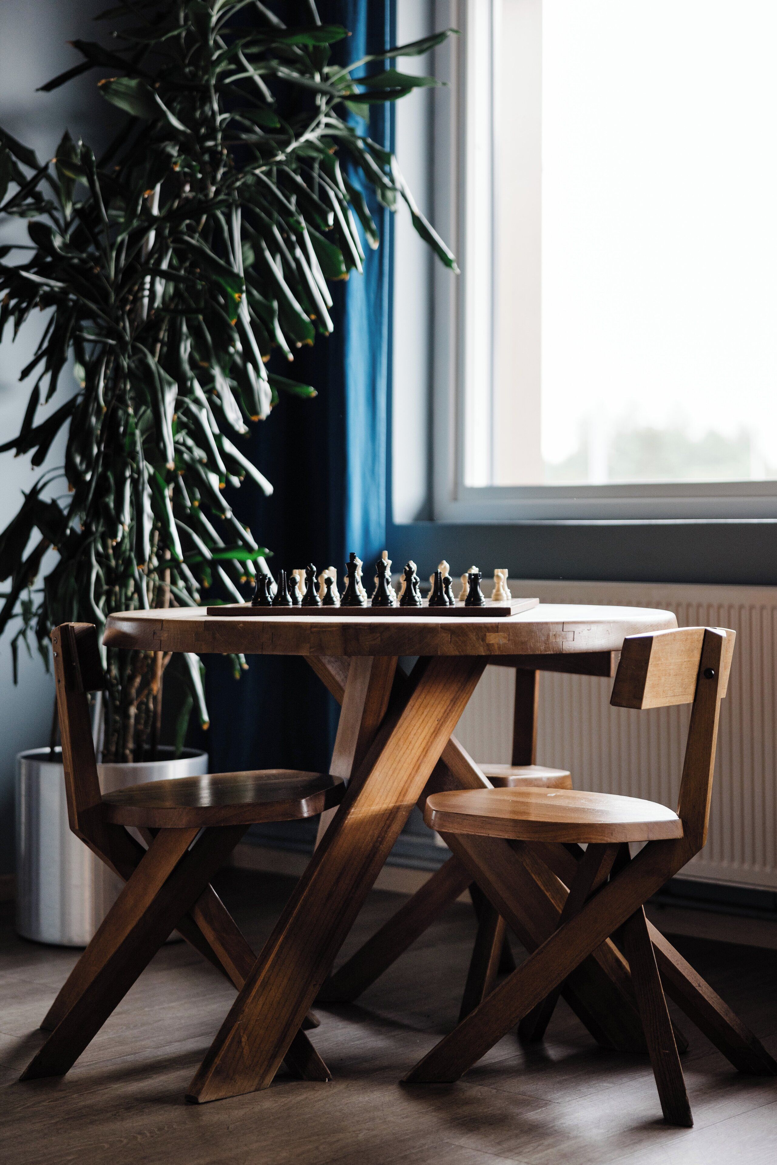 Una mesa de madera para jugar al ajedrez con sillas, una planta en maceta y una ventana al fondo.