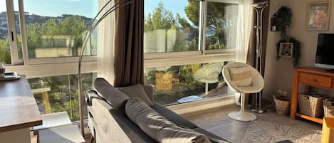 Een moderne woonkamer met een bank, een fauteuil, een televisie en een groot raam met uitzicht op groen.