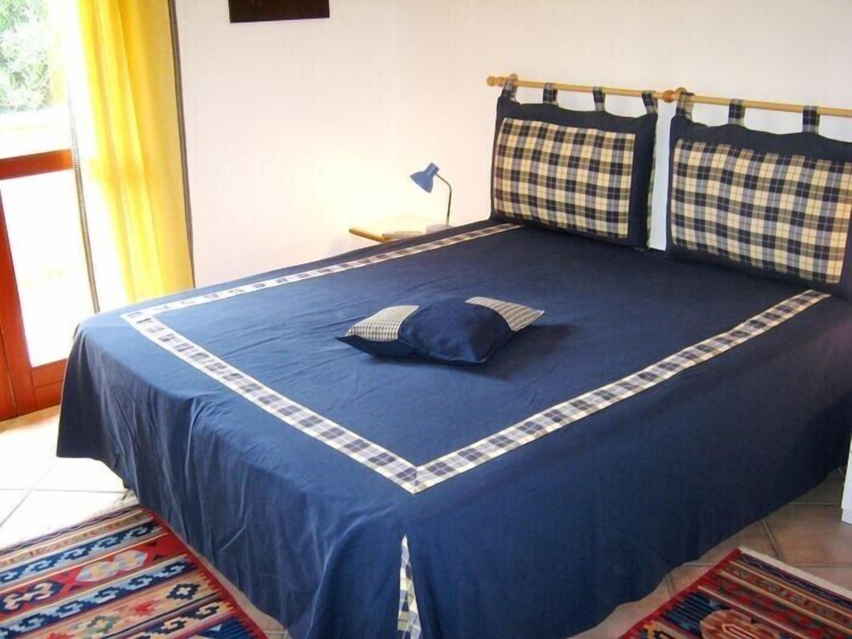 Ein Bett mit blauer Bettdecke und karierten Kissen, einem gemusterten Teppich und einem Holzkopfende.