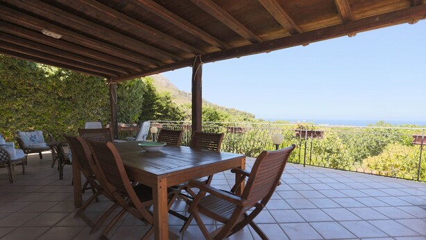 An outdoor patio with a wooden table and chairs, surrounded by a metal fence, overlooking a vineyard and a distant view of the sea.