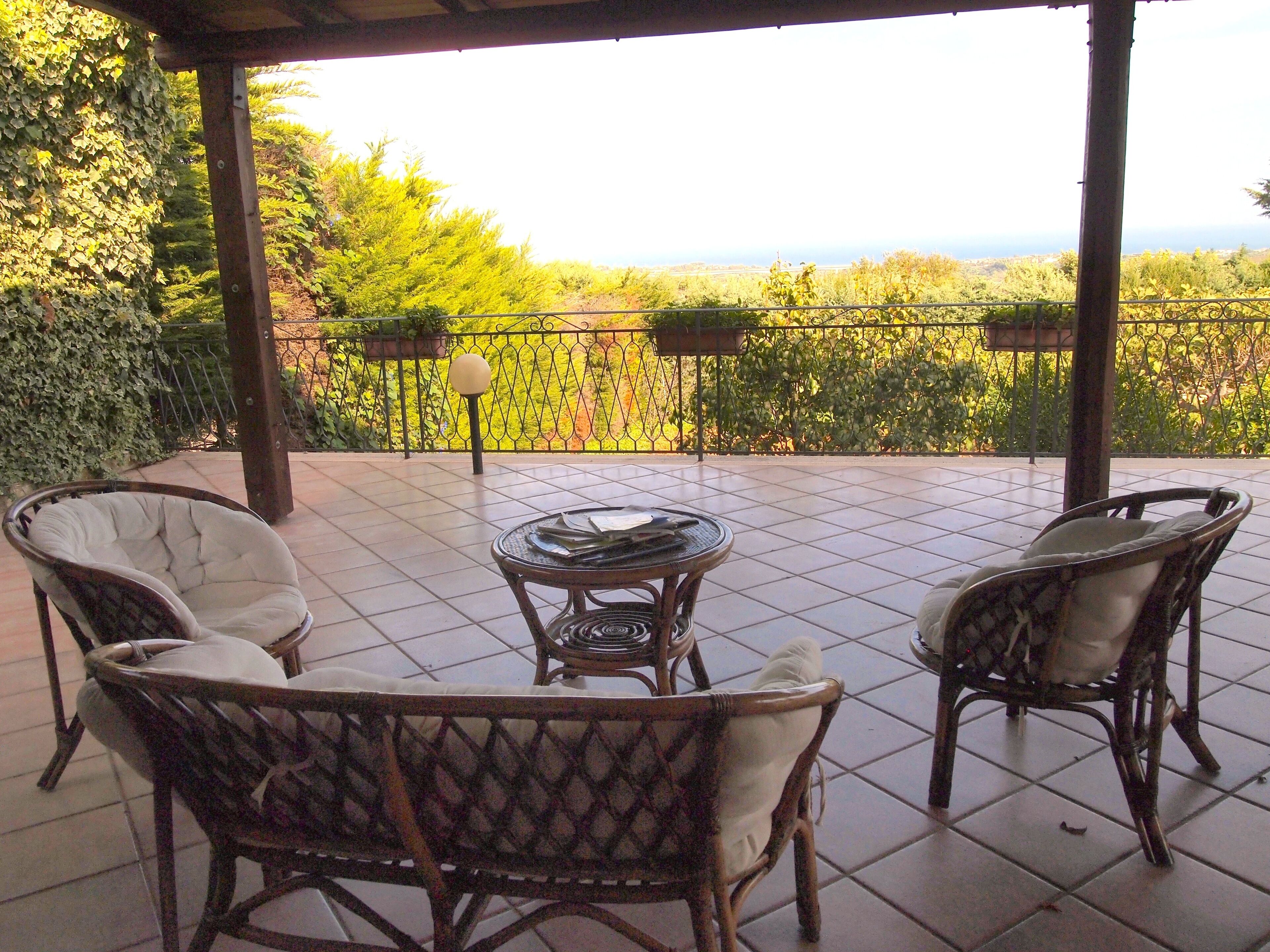 A covered patio with wicker furniture and a table, overlooking a lush garden and distant landscape.