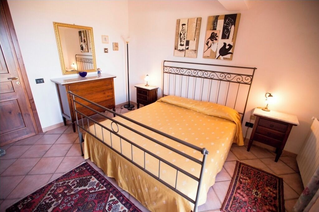 A bedroom with a metal bed frame, a wooden dresser, a bedside table with a lamp, and a patterned rug on the floor.