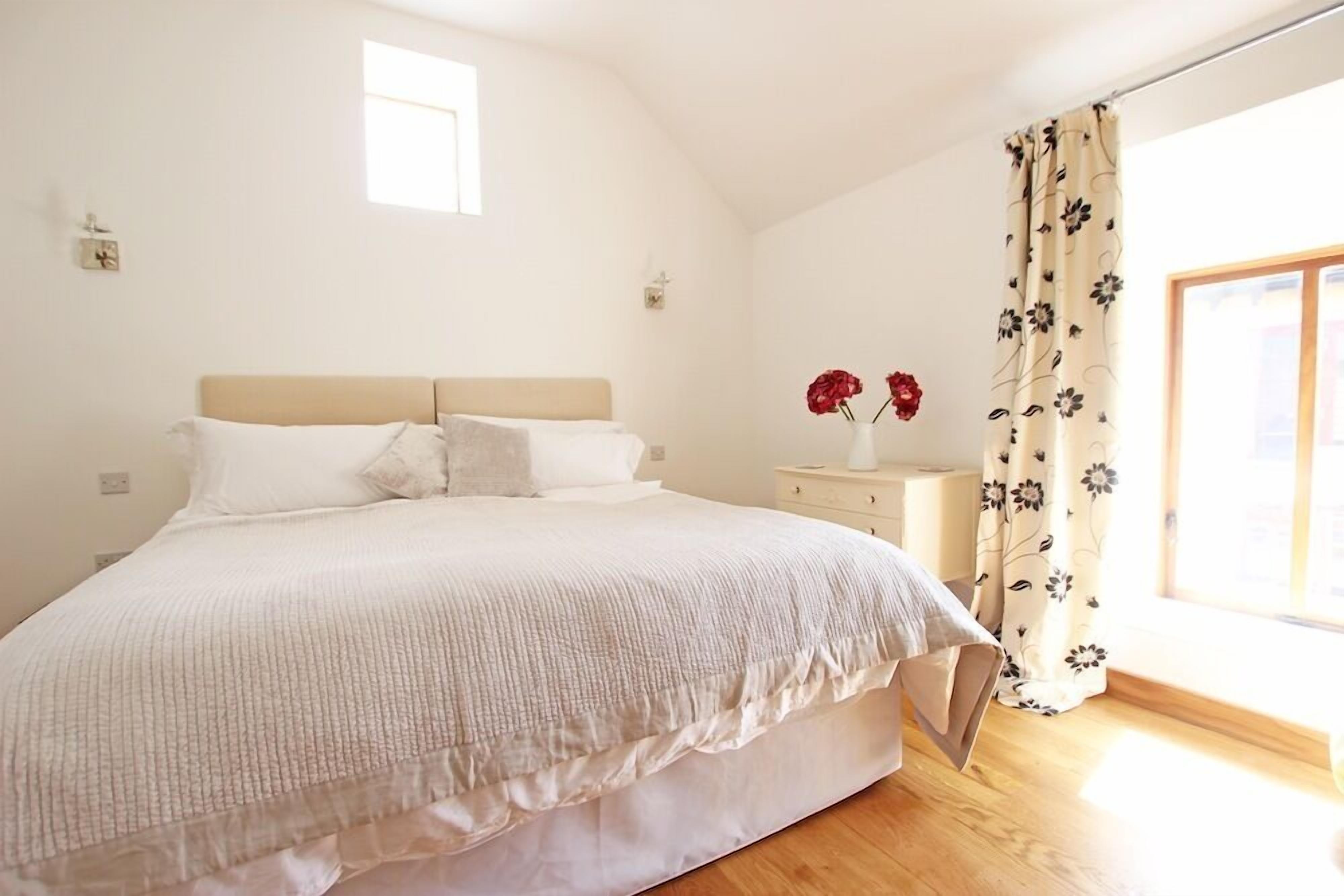Een slaapkamer met een groot bed, een nachtkastje, een vaas met bloemen en een raam met gordijnen.