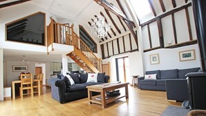A spacious living area with a high ceiling, wooden beams, a staircase, and a fireplace.