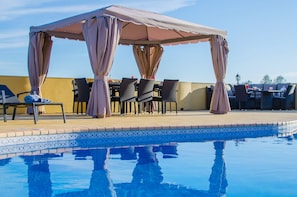 Un espace aménagé au bord de la piscine, comprenant un coin salon couvert, des chaises longues et une piscine.