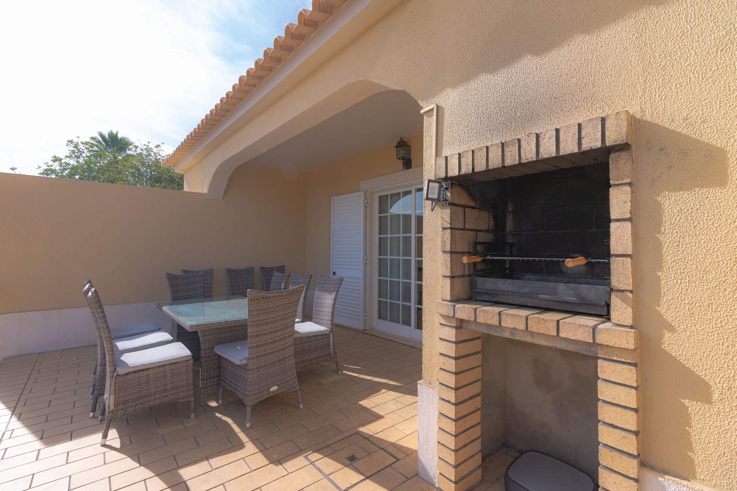 Une terrasse extérieure dotée d’un barbecue en briques, de meubles en osier et d’une porte vitrée donnant accès à un espace abrité.