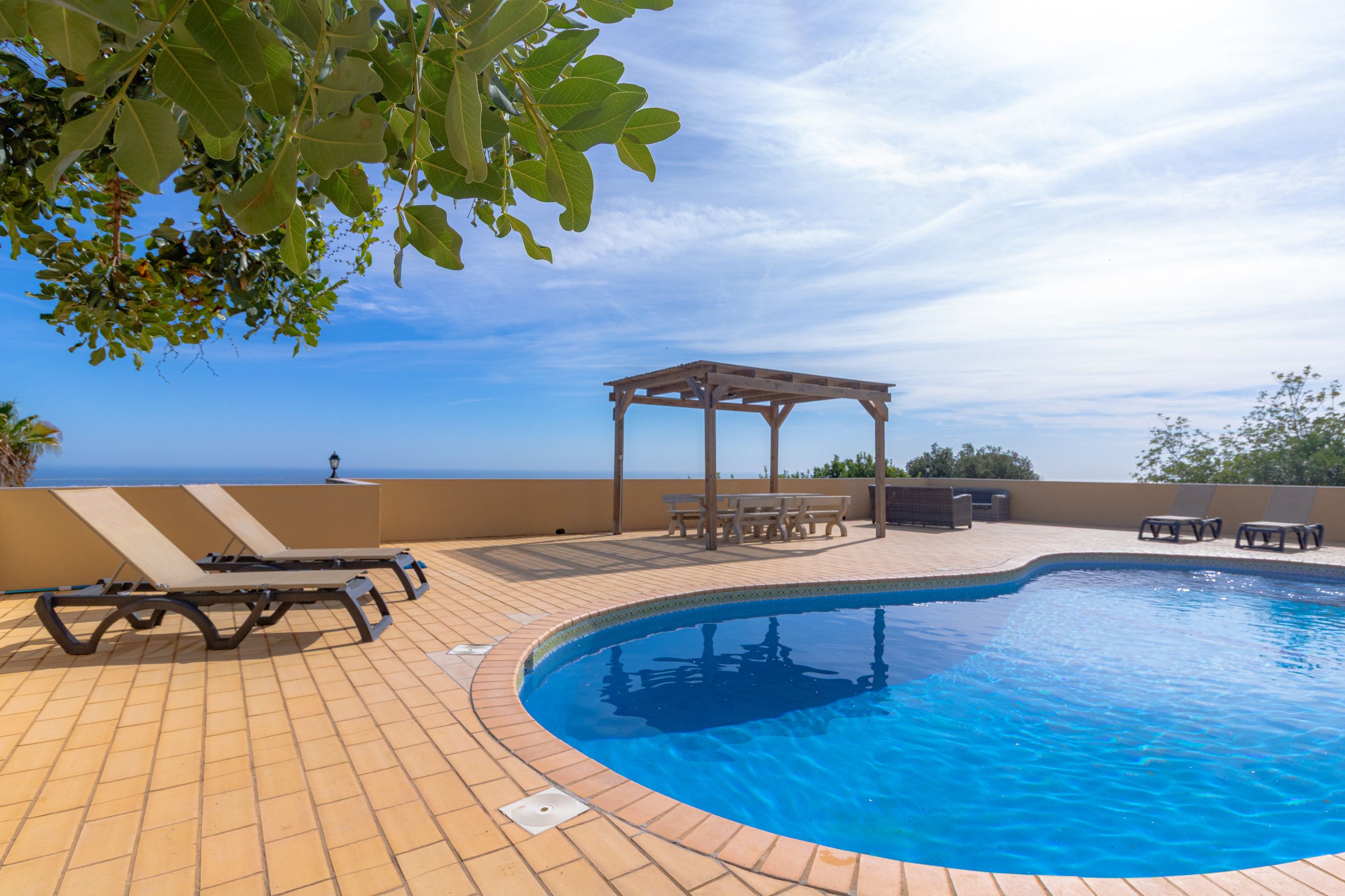 A swimming pool with lounge chairs and a pergola, surrounded by a tiled deck and overlooking a body of water.