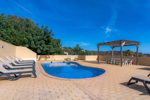 Una piscina con sillones de descanso y una pérgola, rodeada por un patio de ladrillo y follaje.