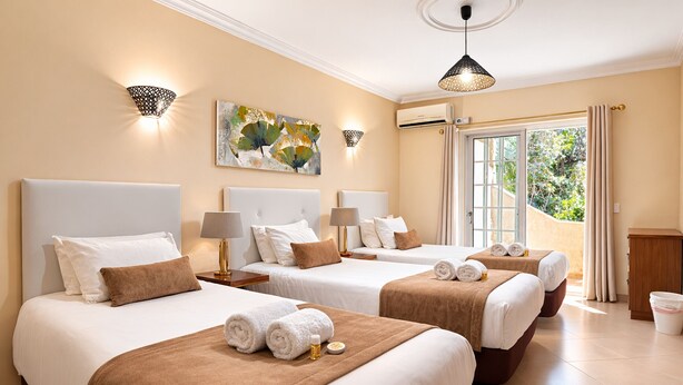 A hotel room with three beds, each with white linens and brown headboards. There are wall-mounted lamps, an air conditioning unit, and a large window with curtains. - Villa Panoramica | 5 Bedrooms | Panoramic Views | Estói (Estoi)