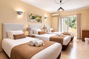 Une chambre d’hôtel avec trois lits, chacun doté de draps blancs et d’une tête de lit marron. On y trouve des lampes fixées au mur, un appareil de climatisation et une grande fenêtre avec des rideaux.