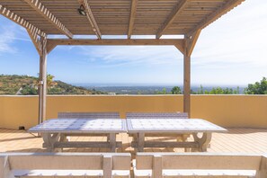 Un patio al aire libre con una pérgola de madera, dos mesas con manteles estampados y vistas a un paisaje costero.