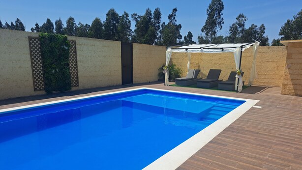 A rectangular swimming pool with a clear blue water surface, surrounded by a wooden deck and a beige wall with a lattice design. - Ideal for families with children, has plenty of room to play. Pets accepted (aveiro-vagos)