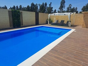 Una piscina rectangular con superficie de agua azul cristalina, rodeada por una terraza de madera y un muro beige con diseño de celosía.