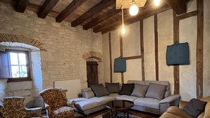 Un salon de style rustique avec des poutres apparentes, un mur en pierre et un grand canapé d’angle. On y trouve deux luminaires suspendus et une petite table au centre.