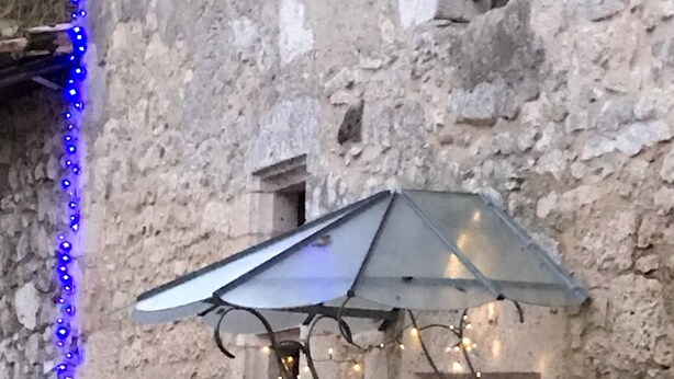 Un bâtiment rustique en pierre, doté d’une structure en métal en forme de parasol, de guirlandes lumineuses bleues et de plantes en pot.