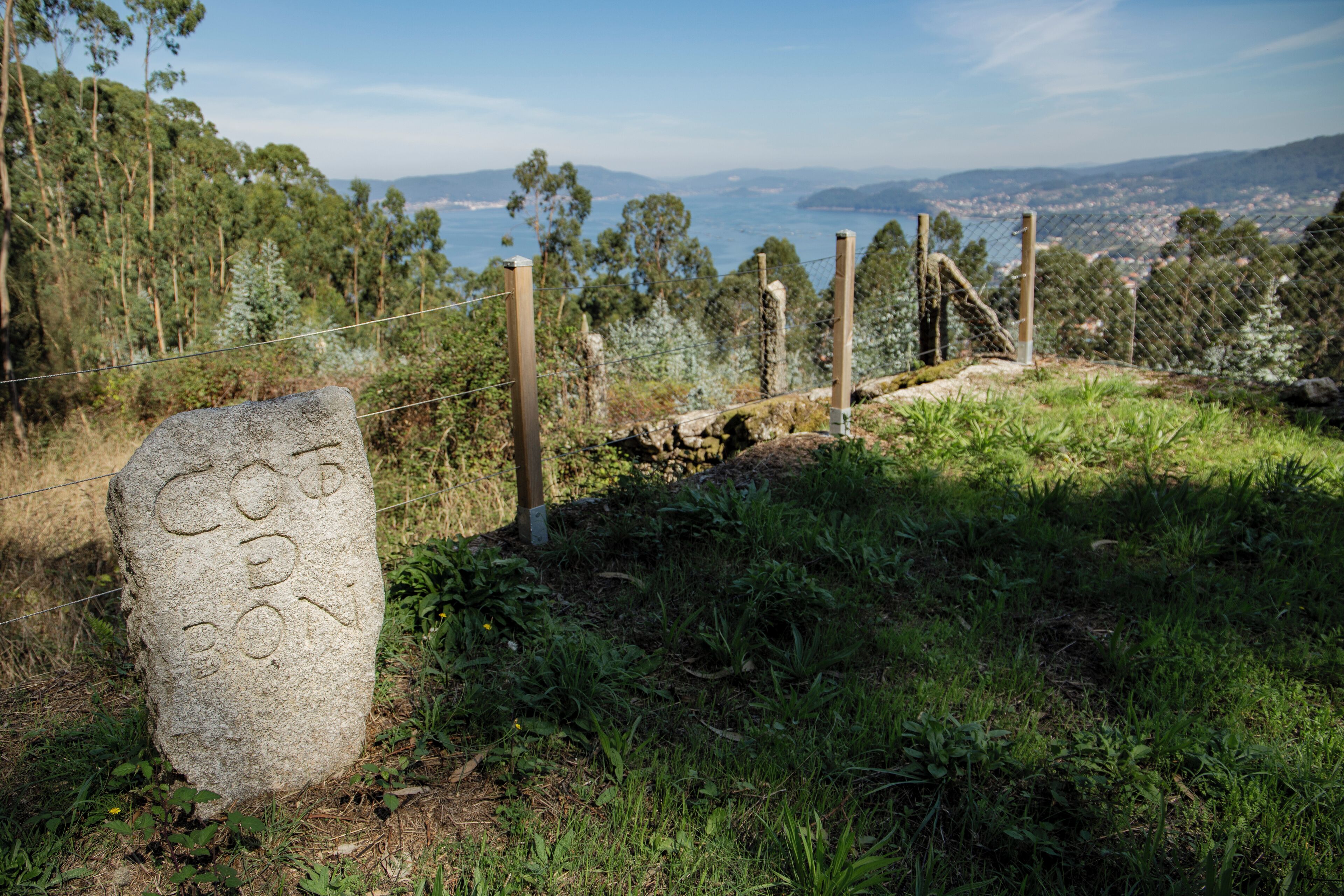 Un monument en pierre portant des inscriptions, une zone délimitée par une clôture et une vue sur une ville côtière.