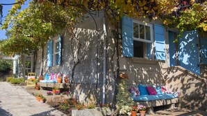 A stone building with blue shutters, a stone bench with colorful cushions, and potted plants.