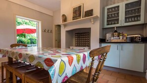 Un comedor con una mesa cubierta con mantel estampado con motivos vegetales, sillas de madera, un ventanal con toldo y una cocina con armarios y electrodomésticos.