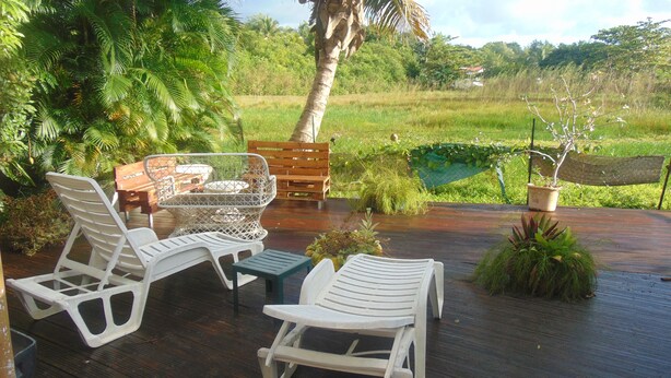 An outdoor seating area with white plastic chairs and a wooden table, surrounded by lush greenery and a view of a rice field. - Caribgites, the Butterfly studio (Sainte-Anne)