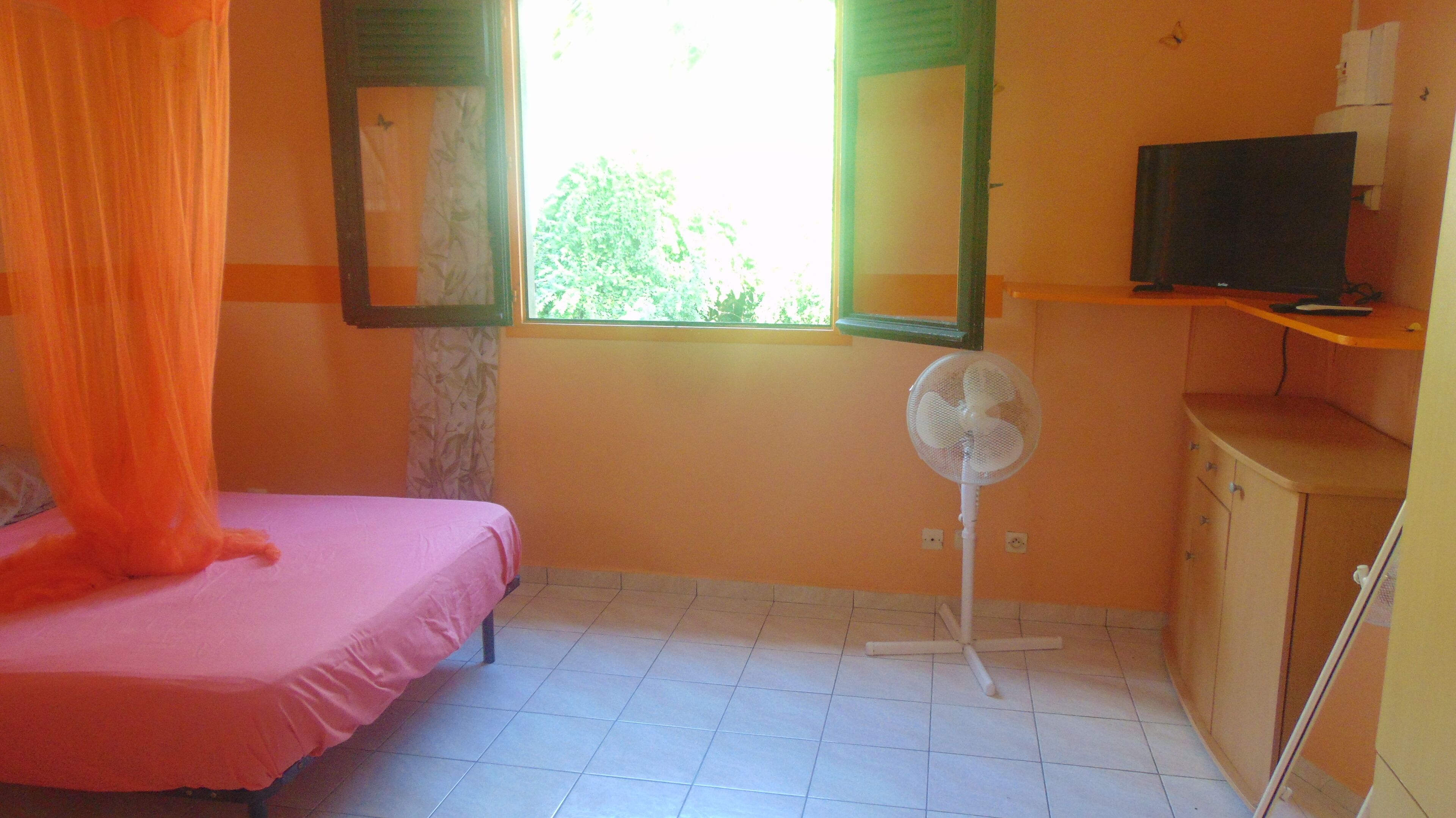A room with a bed, a standing fan, a television on a shelf, and a window with curtains.
