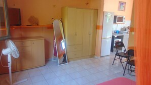 A room with a wooden cabinet, a standing fan, a wardrobe, a chair, and a kitchen area with a refrigerator and microwave. - Caribgites, the Butterfly studio (Sainte-Anne)