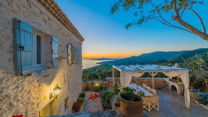A rustic stone building with shutters, a patio with a table and chairs, and a view of the sea and mountains.