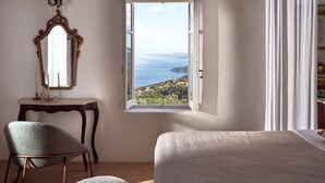 A bedroom with a large window offering a scenic view of the sea and hills.