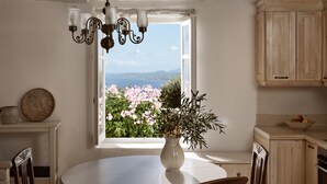 A dining area with a round table, a vase with flowers, a chandelier, and a window with a view of greenery and a distant landscape.
