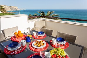 A dining table set with plates, glasses, and a pitcher of orange juice, overlooking a coastal view.