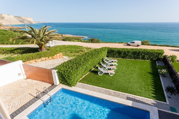 A pool area with a clear view of the sea, a lawn with sun loungers, and a white van parked nearby.