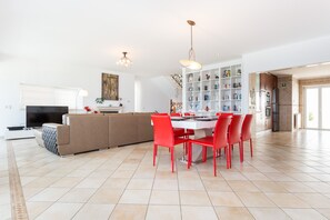 A spacious living area with a large sofa, a dining table with red chairs, and a kitchen with a white cabinet and shelves.