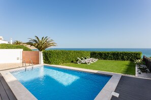 Une piscine avec vue sur la mer, un espace verdoyant avec des chaises longues et une terrasse en bois.