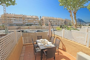 An outdoor patio with a table set for dining, surrounded by wicker chairs, a white fence, and a view of a multi-story building in the background.