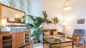 Ein Wohnzimmer mit einer Couch, einem Couchtisch und einem großen Spiegel. Es befinden sich Pflanzen, ein Kaktus als Dekoration und eine Pendelleuchte dort.