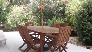 Una mesa de madera redonda con seis sillas bajo un paraguas amarillo, rodeada de exuberante follaje y flores rosadas.