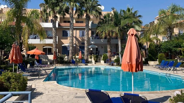 Zona de piscina con sillas de descanso y sombrillas, rodeada de palmeras y edificios con balcones.