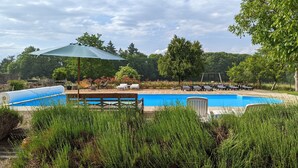Una piscina rodeada de sillas de descanso y una zona cubierta, con árboles y una terraza de madera al fondo.