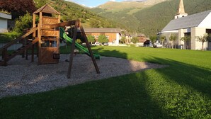 Une aire de jeux avec un toboggan en bois et des balançoires, entourée d’une pelouse et avec des bâtiments en arrière-plan.