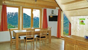 Un coin repas en bois avec une table et des chaises, un téléviseur fixé au mur et une vue sur les montagnes à travers de grandes fenêtres.
