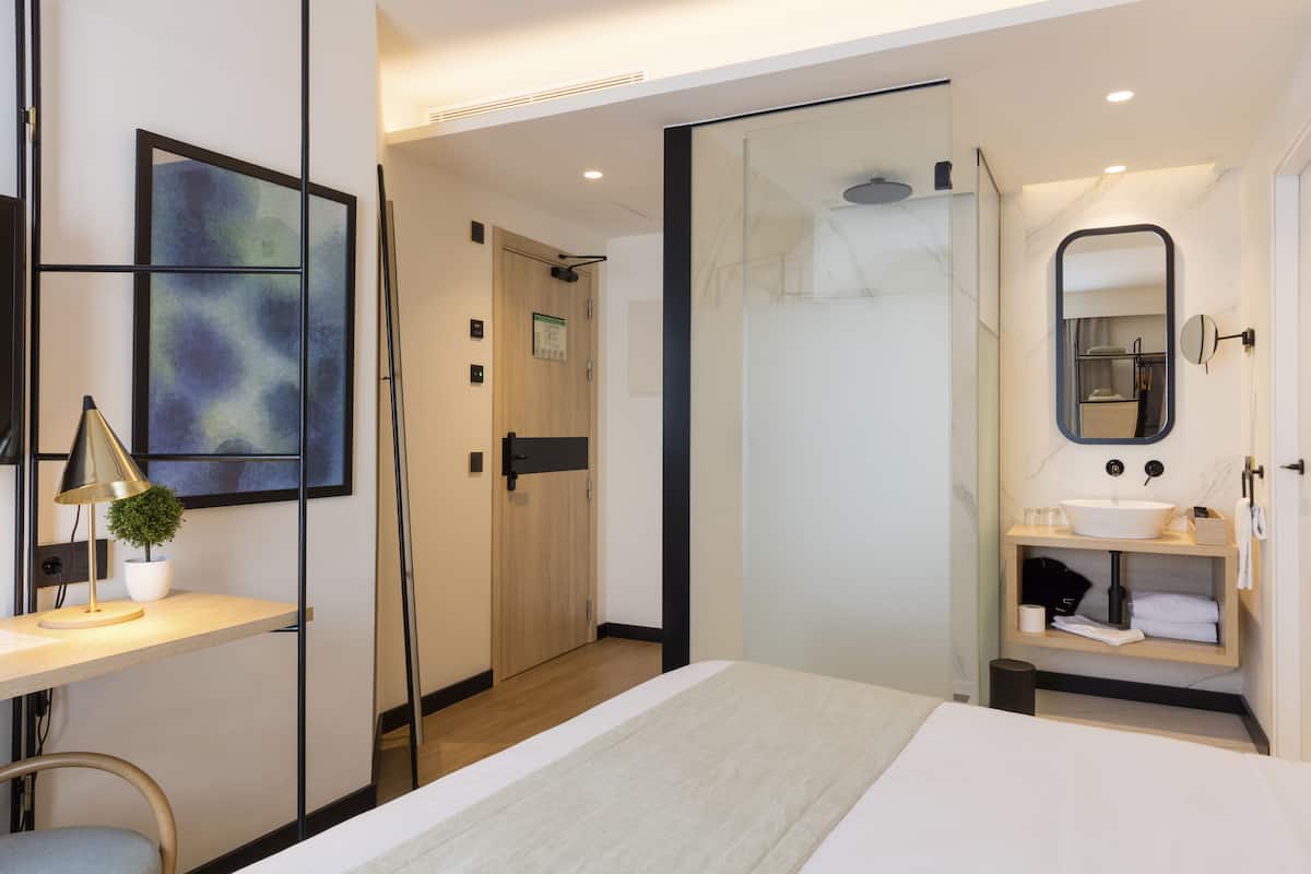 Una habitación de hotel moderna con una cama, un escritorio con lámpara, una planta en maceta, cabina de ducha de vidrio, baño con lavabo y espejo, y una puerta que da a otra habitación.