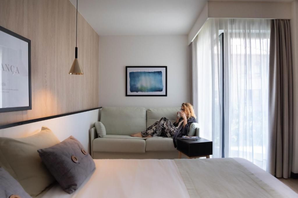 Habitación de hotel con cama, sofá, mesita auxiliar, un cuadro enmarco y una ventana con cortinas.