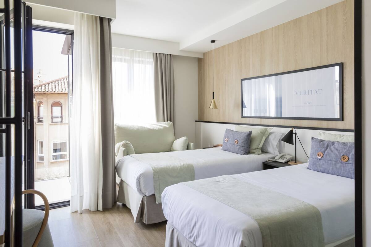 Habitación de hotel con dos camas, un escritorio, una silla, una ventana con cortinas, una obra de arte en la pared y vistas a edificios.