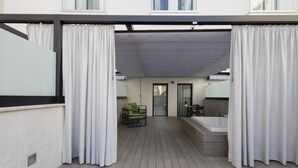 A modern outdoor balcony with a wooden floor, a seating area with a table and chairs, and a large bathtub.