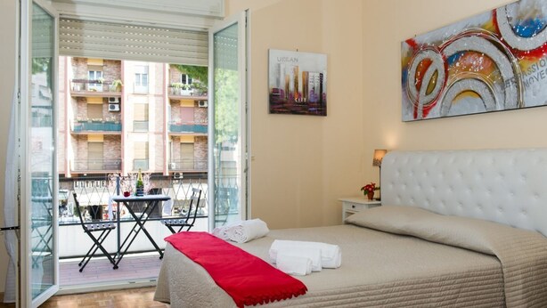 A hotel room with a bed, a red throw blanket, a cityscape painting, and a balcony with a table and chairs. - House Gregorio, large and comfortable apartment (Roma)