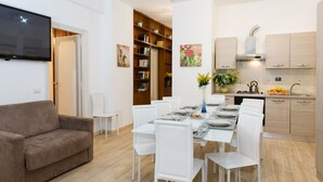 A modern living room with a brown sofa, white dining table set for four, and a kitchen with wooden cabinets and a built-in oven. - House Gregorio, large and comfortable apartment (Roma)