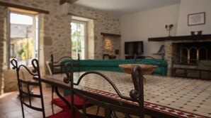 Un salon chaleureux avec un mur en pierre, une cheminée, un téléviseur et un coin repas agrémenté d’une nappe à motifs.