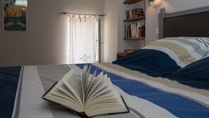 Une chambre à coucher avec un lit, un livre ouvert, une fenêtre avec des rideaux, une bibliothèque et une lampe fixée au mur.